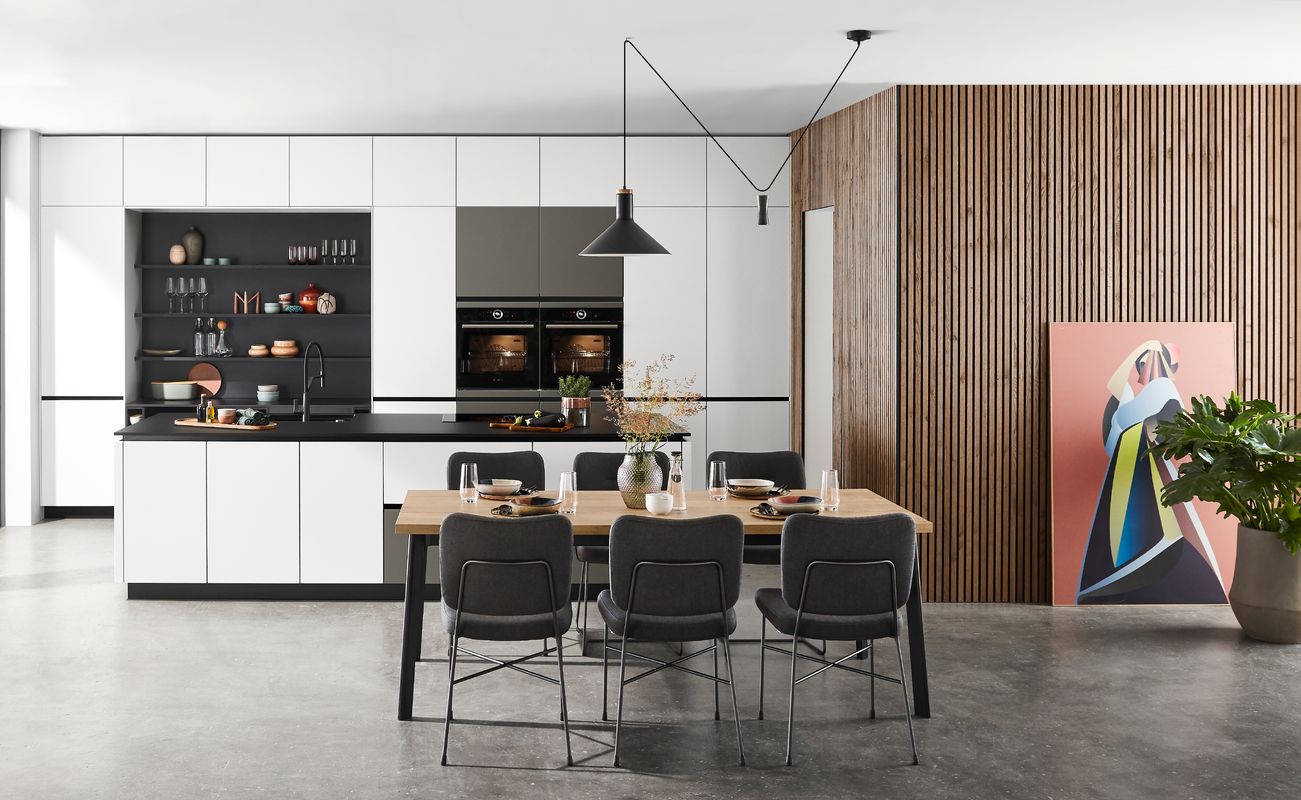 Moderne Küche mit Essbereich, Holzverkleidung und großen Fenstern. Ein Tisch mit Stühlen steht im Vordergrund, Kunstwerk an der Wand.